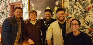 Lab photo showing 5 people standing next to a bar in front of a painting and a wall with floral wall paper